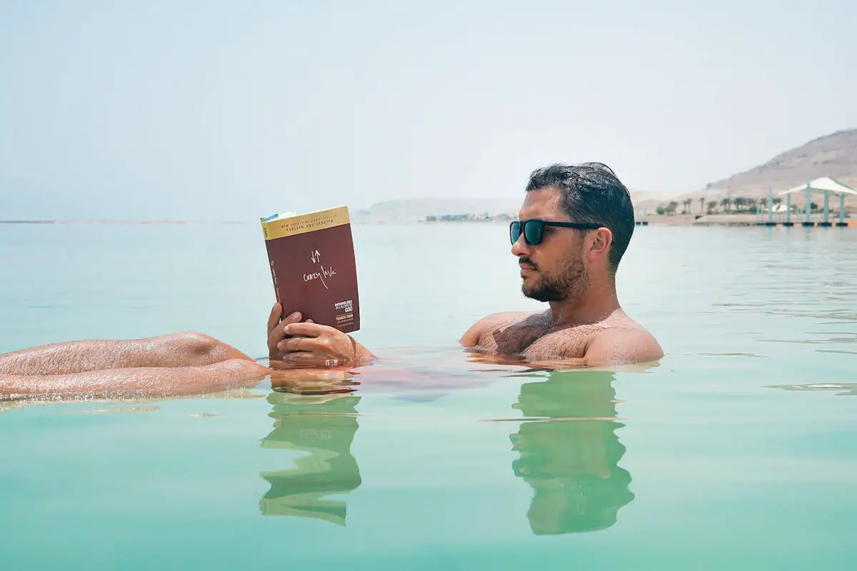man wearing sunglasses reading book on body of water
